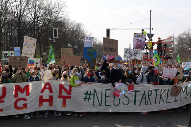 Eine große Gruppe von Menschen marschiert auf einer Straße, hält ein 'Menschenrechte'-Schild und verschiedene Plakate, wobei einige Masken tragen; Bäume, Laternenmasten und ein klarer blauer Himmel sind im Hintergrund zu sehen.