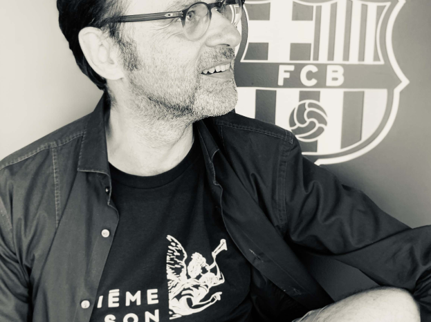 Ein Mann mit Brille und schwarzem Hemd lächelt, während er vor dem FC Barcelona-Logo an einer Wand sitzt, auf einem Schwarz-Weiß-Foto.