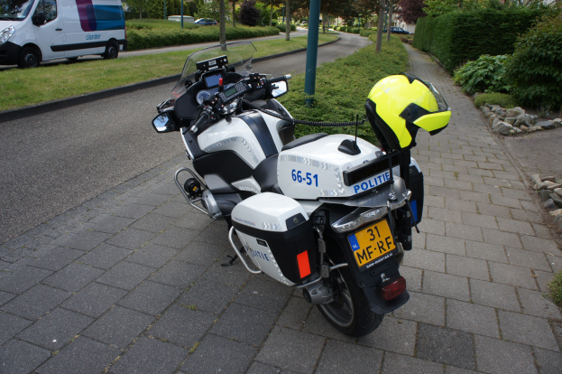 Ein polizeiliches Motorrad am Straßenrand mit einem Helm darauf, umgeben von Bäumen, Pfählen, Gebäuden, Pflanzen, Gras, Steinen und anderen Fahrzeugen im Hintergrund.