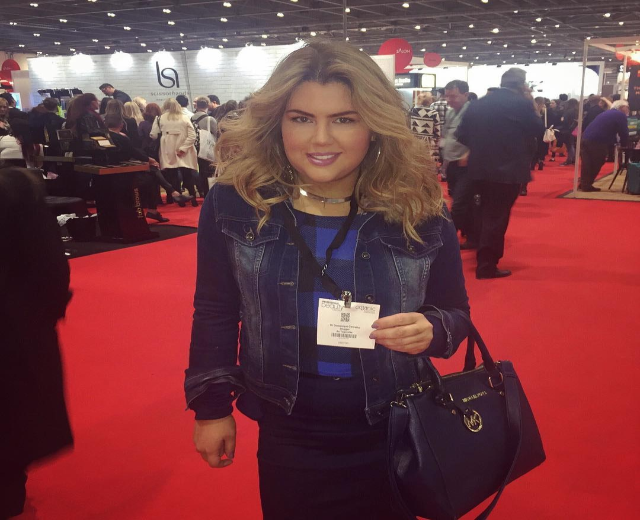 Frau auf einem roten Teppich an einer Modenschau, die lächelt und eine Handtasche hält, mit Menschen, Lichtern und Plakaten im Hintergrund.