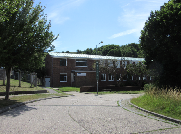 Ein Schulgebäude mit Fenstern, einem Straßenpfahl, einem Schild, einer Baumgruppe, Gras, einem Zaun und einem bewölkten Himmel, das inmitten eines Parks umgeben von saftigem Gras liegt.