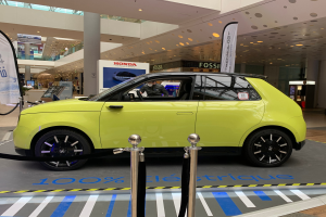 Ein gelbes wasserstoffbetriebenes Elektroauto steht vor einem Gebäude, umgeben von Absperrpollern mit Seilen, Bannern und Schildern, mit einer Person im Auto und einer Gruppe von Menschen in der Nähe, unter Deckenleuchten und einer Decke mit Deckenventilatoren.