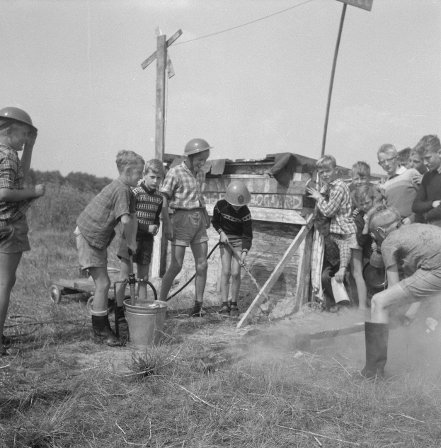 Schwarz-weißes Bild von Kindern in Helmen, die um einen Hydranten in einem grasbewachsenen Feld stehen, einige halten Rohre, mit einem Eimer, einer Karre, Stangen mit Drähten und einer Tafel mit Text in der Nähe, Bäume im Hintergrund unter einem klaren Himmel.