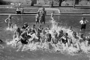 Schwarzes und weißes Bild von Kindern, die in einem Swimmingpool spielen, mit Bäumen und Gebäuden im Hintergrund.