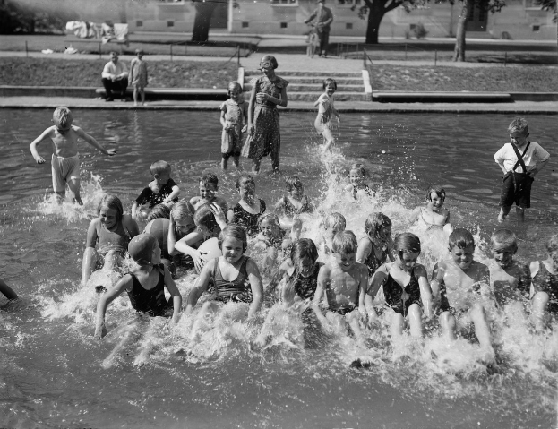 Schwarzes und weißes Bild von Kindern, die in einem Swimmingpool spielen, mit Bäumen und Gebäuden im Hintergrund.
