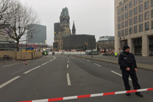 Polizist in schwarzer Uniform mit Mütze auf einer Berliner Straße umgeben von Absperrbaken, Fahrrädern, Laternenmasten, Bäumen und Gebäuden, mit einem Kirchturm im Hintergrund bei klarem blauem Himmel.