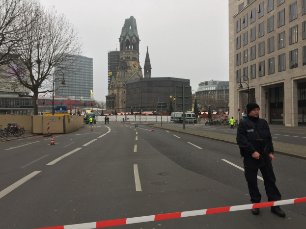 Polizist in schwarzer Uniform mit Mütze auf einer Berliner Straße umgeben von Absperrbaken, Fahrrädern, Laternenmasten, Bäumen und Gebäuden, mit einem Kirchturm im Hintergrund bei klarem blauem Himmel.