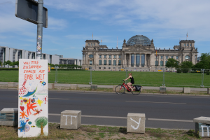 Eine Person in einem schwarzen Kleid fährt mit dem Fahrrad eine Straße vor dem Reichstaggebäude in Berlin, Deutschland, entlang, mit Gebäuden, Bäumen und einigen Menschen im Hintergrund.