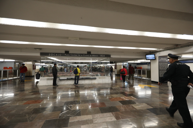 Eine Gruppe von Menschen, die durch eine belebte U-Bahn-Station gehen, mit einem Polizeibeamten im Vordergrund, überkopfschilder mit Text und Bildschirmen sowie verschiedene strukturelle Elemente wie Geländer.