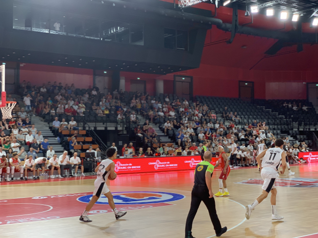 Eine Gruppe von Menschen, die Basketball in einer Turnhalle mit einem Netz auf der linken Seite spielen, Zuschauer im Hintergrund und Deckenbeleuchtung.