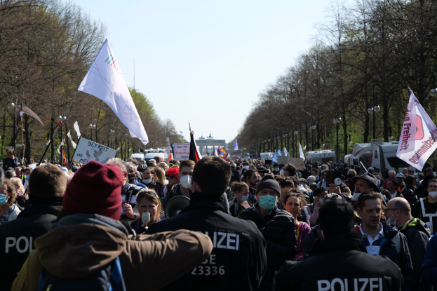 Eine große Gruppe von Menschen steht vor einer Menge von Polizeibeamten, einige tragen Mützen und Masken, während sie bei einer Demonstration in Berlin, Deutschland, mit Schildern, Fahnen, Laternenpfählen, Bäumen, Fahrzeugen, einem Gebäude und dem Himmel im Hintergrund stehen.