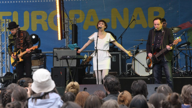 Vier Musiker auf der Bühne: Eine Frau in einem weißen Kleid singt in der Mitte, flankiert von zwei Männern mit Gitarren, mit einem Publikum unten und Lautsprechern an einer Wand im Hintergrund.