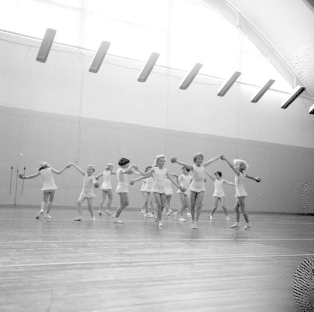 Schwarzes und weißes Bild von jungen Mädchen in einem Tanzstudio, die sich an den Händen halten und tanzen, mit einem Ball auf der rechten Seite, Deckenlampen und einem wandmontierten Objekt im Hintergrund.
