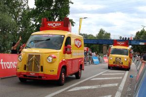 Zwei gelbe und rote Lkw fahren eine von Menschen gesäumte Straße mit Barrikaden und Schildern entlang, im Hintergrund eine Brücke, Bäume, Laternen, Drähte und ein bewölkter Himmel.