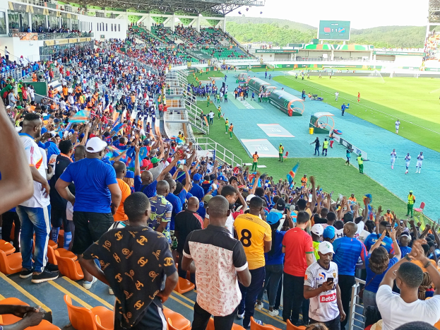 Eine große Menschenmenge versammelt sich vor einem eingezäunten Fußballfeld, einige sitzen und andere stehen, mit Blick auf Hügel unter einem klaren blauen Himmel während der Afrika-Cup 2019.