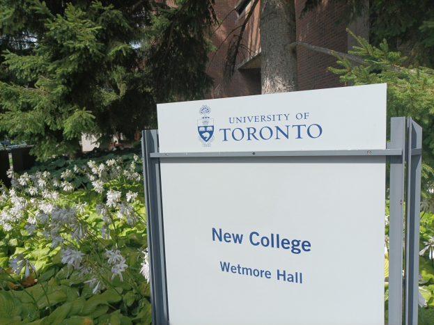 Außenansicht des neuen Colleges der Universität Toronto an der Wetmore Hall mit einem Schild vor dem Gebäude, umgeben von grünem Gras, bunten Blumen, hohen Bäumen und einem klaren blauen Himmel.