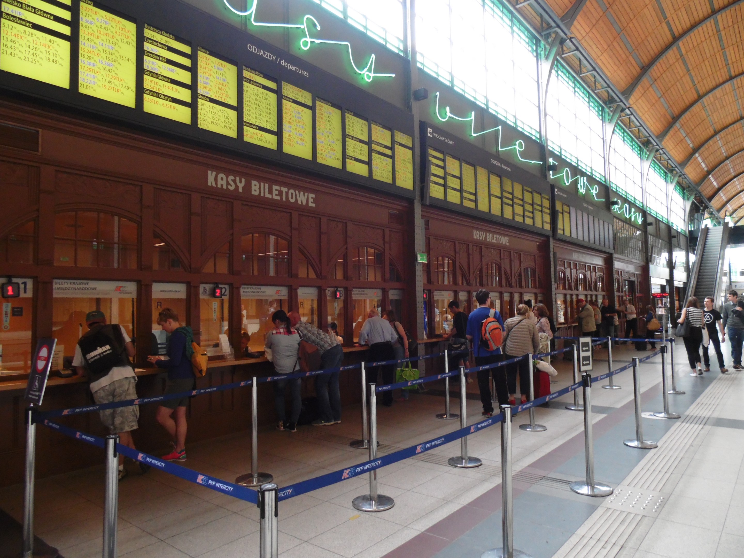 Eine Gruppe von Menschen steht an einem Bahnhof in der Schlange an einem Ticketschalter, einige tragen Taschen, unter Deckenlampen, in der Nähe von Pfosten mit Bändern und Informationsschildern, mit Stufen und Gelöndern, die zu einem Gebäude im Hintergrund führen.