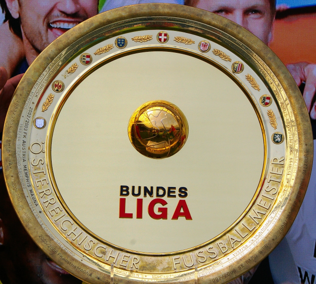 Goldener Schild mit "Bundesliga"-Text in den Händen eines Mannes, mit einem Banner im Hintergrund mit drei Personen.