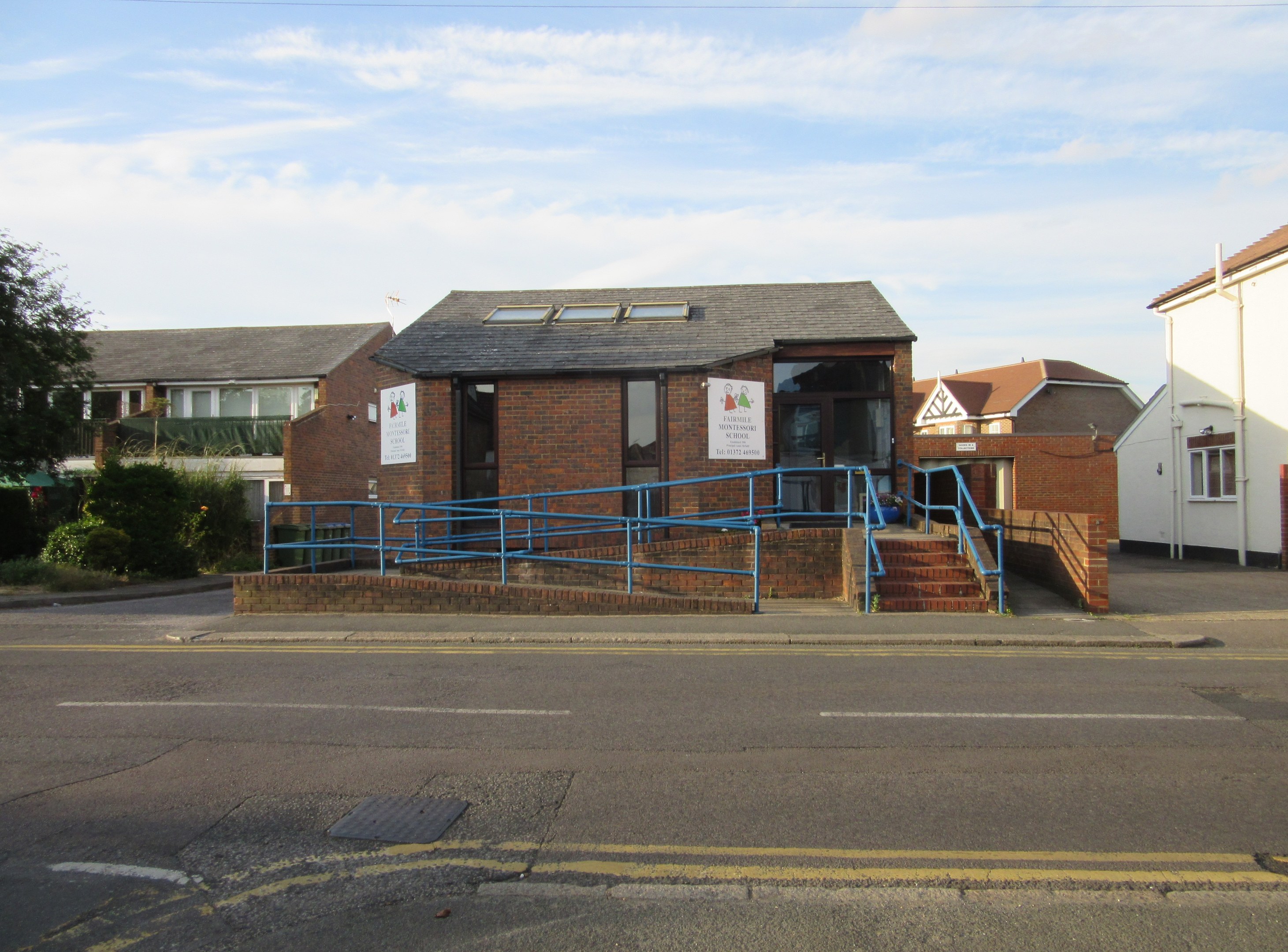 Ein Schulgebäude mit blauer Begrenzung, umgeben von Häusern, Treppen mit Geländern, Texttafeln, Pflanzen, Bäumen und einem bewölkten Himmel.