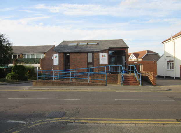 Ein Schulgebäude mit blauer Begrenzung, umgeben von Häusern, Treppen mit Geländern, Texttafeln, Pflanzen, Bäumen und einem bewölkten Himmel.