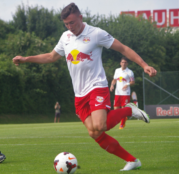 Ein professioneller Fußballspieler, der einen Ball auf einem grünen Feld mit Bäumen und einem klaren blauen Himmel im Hintergrund kickt, wobei ein 'RB Leipzig'-Schild sichtbar ist.