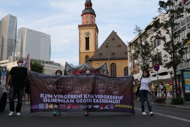 Gruppe von Menschen in Masken, die eine Protestfahne schwenken und eine Straße entlanggehen, mit einem geparkten Auto auf der linken Seite und Gebäuden, Bäumen und einem Kirchturm im Hintergrund unter einem klaren blauen Himmel.