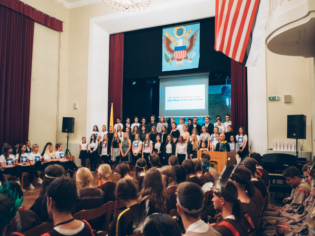 Eine Gruppe von Menschen steht an einem Podium mit einem Mikrofon, flankiert von Fahnen und Lautsprechern, vor einem sitzenden Publikum während eines kulturellen Ereignisses, mit einem Bildschirm, Vorhängen und einem Kronleuchter im Hintergrund.