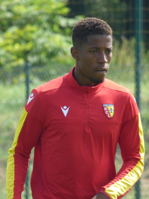 Ein junger Mann in einem roten und gelben Fußballtrikot steht auf einem Feld und hält die Umrandung mit entschlossener Miene fest, Bäume sind im Hintergrund zu sehen.