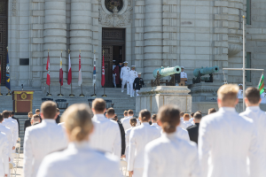 Gruppe von Menschen in weißen Marineuniformen vor einem Gebäude mit Säulen und Treppe während einer Abschlusszeremonie mit Fahnen, einem Podium und Kanonen im Hintergrund.