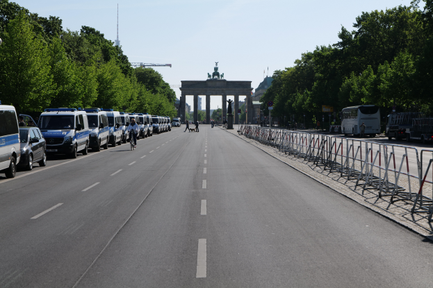 Eine Reihe von Polizeiwagen, die auf der Seite einer Straße vor dem Brandenburger Tor in Berlin, Deutschland, geparkt sind, mit Menschen, die Fahrräder fahren und auf der Straße stehen, Absperrungen und Bäume, die die Seiten säumen, und einem Bogen mit Statuen im Hintergrund.