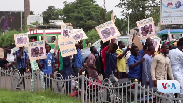 Eine Gruppe von Menschen hält Schilder in Händen und steht vor einem Zaun, umgeben von Fahrzeugen, Bäumen, Masten, Werbetafeln und einem Bildschirm, mit Gras unten und dem Himmel im Hintergrund, scheinbar protestieren sie gegen eine Regierungsentscheidung.