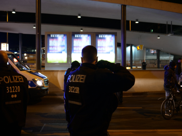 Gruppe von Polizeibeamten steht vor einem Gebäude in der Nacht mit einem geparkten Auto auf der linken Seite und einem Radfahrer auf der rechten Seite, beleuchtet von Bildschirmen und Lichtern im Hintergrund.