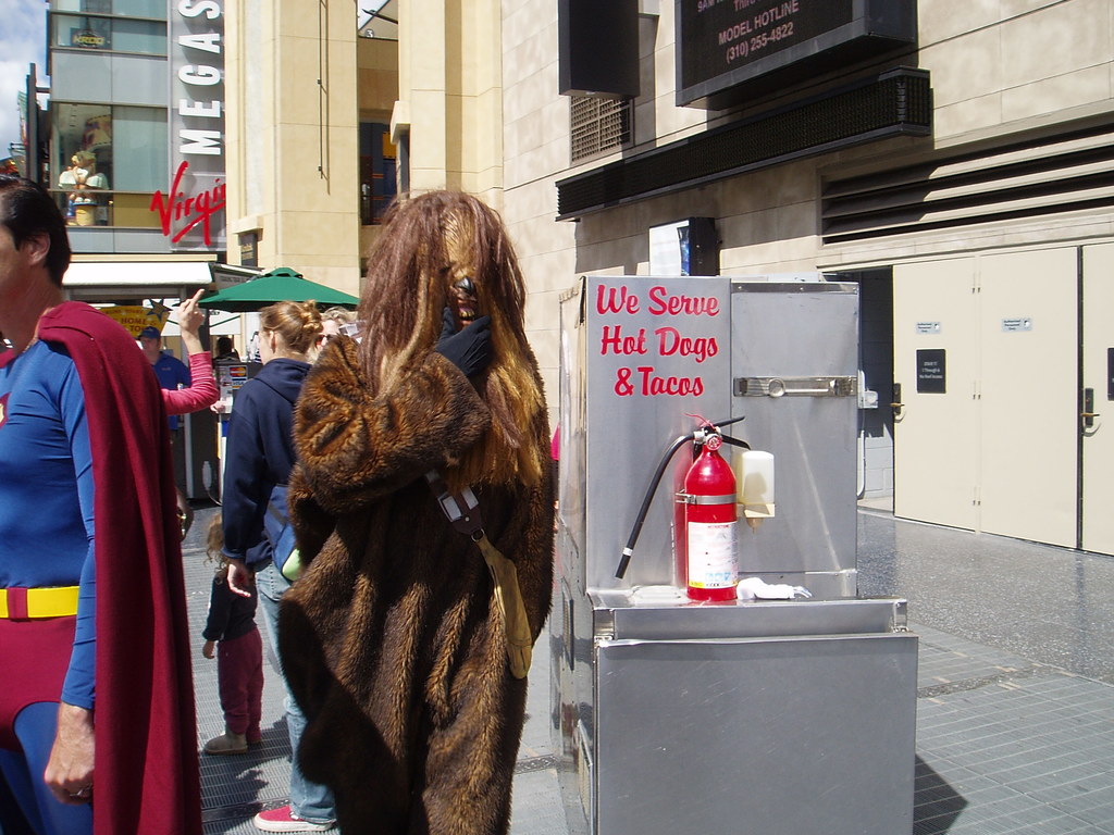 Eine Gruppe von Menschen, einige in Kostümen, versammelte sich vor einem Gebäude mit sichtbarer Beschriftung und Türen, mit einer Box und einem Feuerlöscher in der Nähe.