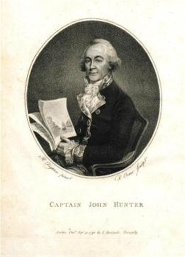 Captain John Hunter, der eine blaue Anzug mit einem weißen Hemd und einer roten Krawatte trägt, sitzt in einem Stuhl und liest ein Buch, mit einem ernsten Gesichtsausdruck. Der Text "Captain John Hunter" ist unten auf dem Bild zu sehen.