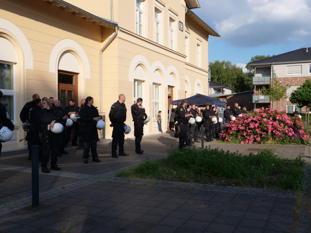 Eine Gruppe von Polizeibeamten vor einem Gebäude mit Fenstern und Türen, die Helme in den Händen halten, in der Nähe eines Zeltes, umgeben von Pflanzen, Blumen, Gras, einem Laternenpfahl, Bäumen und einem bewölkten Himmel.