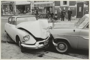Schwarz-weißes Foto von zwei beschädigten Autos am Straßenrand mit Menschen und Fahrrädern in der Nähe und Gebäuden im Hintergrund.