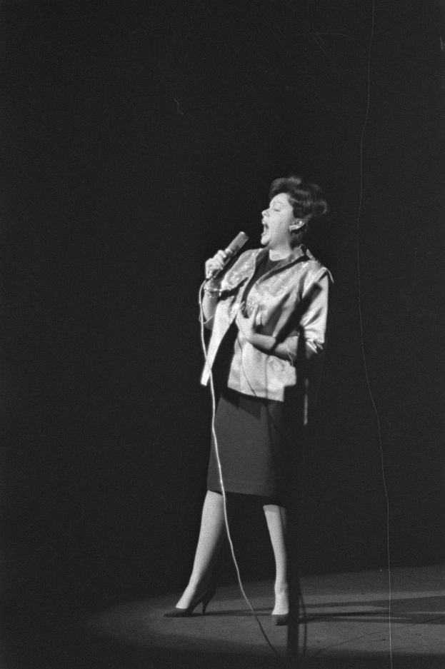 Eine Frau in einem schwarz-weißen Outfit aus den 1950er Jahren singt auf einer von einem Scheinwerfer beleuchteten Bühne und lächelt dabei.