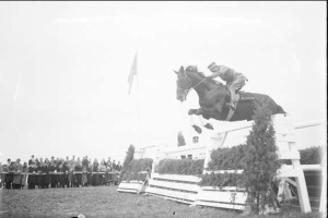 Schwarz-weiß-Foto eines Pferdes und Reiters, die über ein Hindernis springen, bei den Royal Ascot Horse Trials 1953 mit Zuschauern, einer Fahne und Grün im Hintergrund.