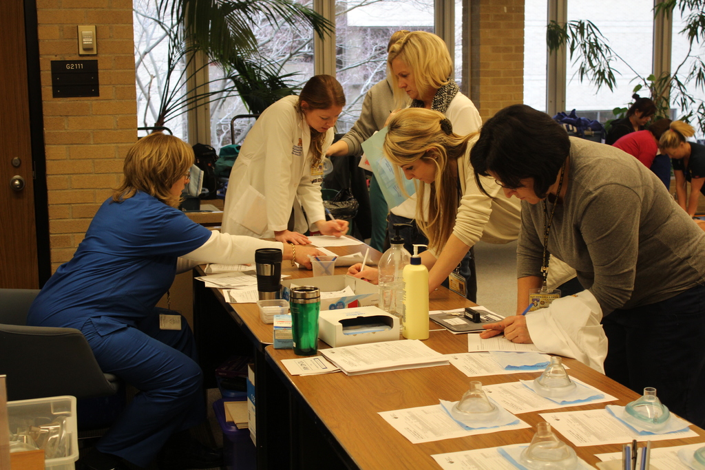 Ein Raum mit Menschen, die neben einem Schreibtisch mit Papieren, Büchern und Flaschen stehen, einer Frau in einem blauen Kleid in der Ecke und großen Glasfenstern mit Pflanzen im Hintergrund.