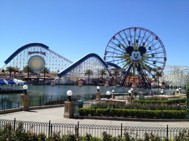 Disneylands California Adventure Mit Einer Riesenrad, Achterbahn, Grünflächen, Laternen, Einem Gewässer Im Vordergrund Und Einem Hellblauen Himmel.