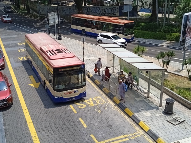 Eine Stadtstraße mit Autos und Bussen, eine Bushaltestelle mit Menschen und Gebäude im Hintergrund.