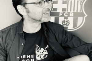Ein Mann in einer Brille und einem schwarzen Hemd lächelt, während er vor einem schwarz-weißen FC Barcelona-Logo sitzt.