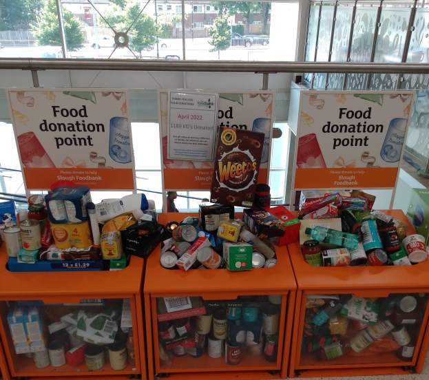 Orangene Schränke voller Lebensmittelspenden, darunter Flaschen, Dosen und Kartons, mit Informationsschildern und einem Blick durch Glasfenster auf Fahrzeuge, Bäume, Gebäude und Himmel.