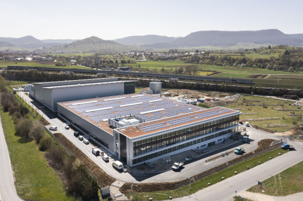 Luftaufnahme eines großen Gebäudes mit Solarmodulen auf dem Dach, umgeben von Fahrzeugen, Bäumen, Gras und Masten, vor Bergen und einem klaren blauen Himmel, identifiziert als Deutschlands erste solarbetriebene Fabrik.