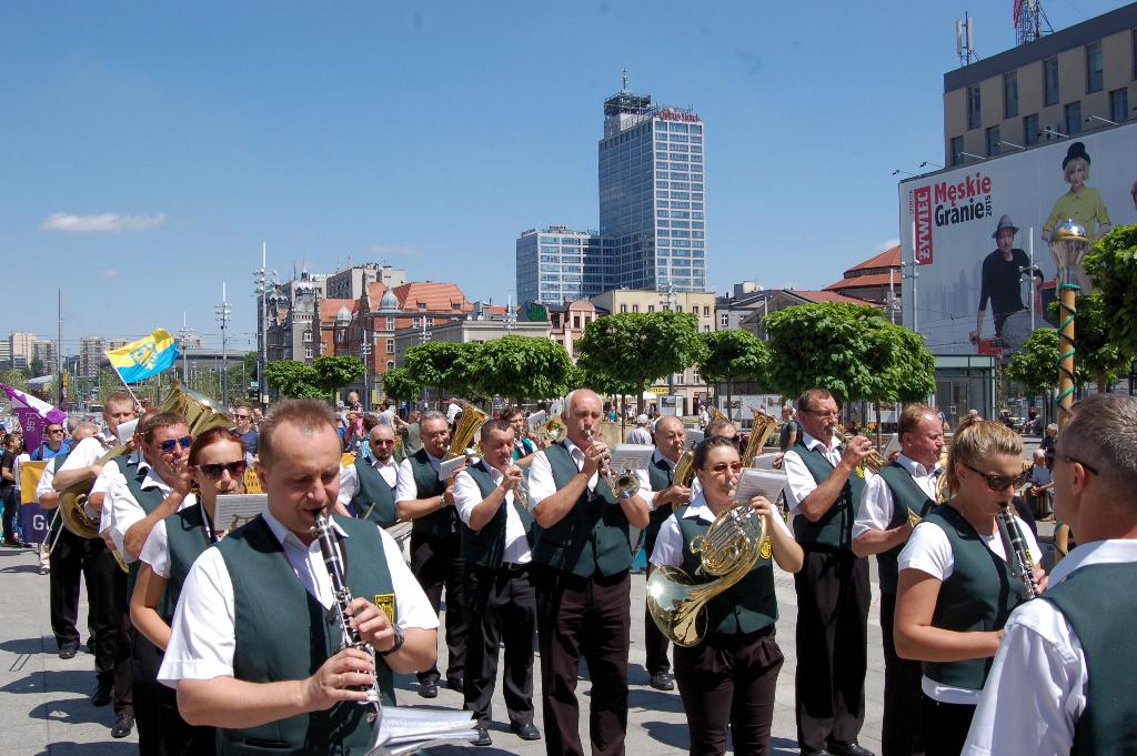 Eine Gruppe von Menschen in einheitlichen Kostümen, die verschiedene Musikinstrumente wie Klarinetten, Tuben und eine French Horn im Freien spielen, mit Bäumen, Polen, Fahnen, einem Banner mit Text und Bildern, einer Lampe, Gebäuden und anderen Objekten im Hintergrund.