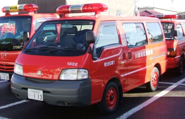 Drei rote Rettungswagen mit weißen und schwarzen Akzenten nebeneinander auf einem Parkplatz geparkt, mit Gebäuden, Polen und einem klaren blauen Himmel im Hintergrund.