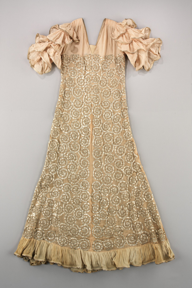 Ein cremefarbenes Kleid mit goldener Stickerei und Rüschen an einer weißen Oberfläche, das mit detaillierten Designs und Mustern verziert ist.