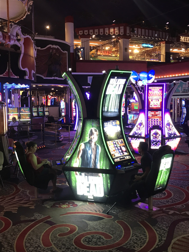 Gruppe von Menschen, die Spielautomaten in einem abgedunkelten Casino bei Nacht mit beleuchteten Gebäuden im Hintergrund spielen.