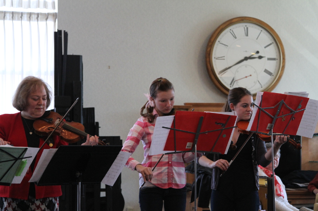 Drei Frauen auf der Bühne mit Geigen spielend, mit Notenpulten und Noten davor, während ein Zuschauer zuhört, mit einer Uhr an der Wand und einem weißen Vorhang im Hintergrund.