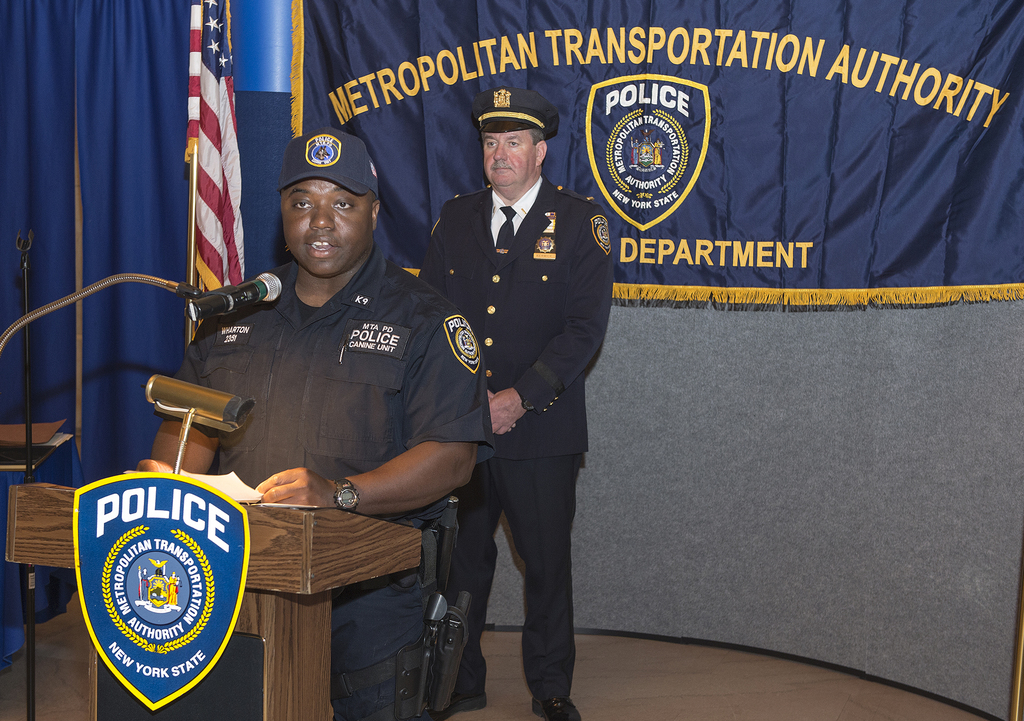 Zwei Polizeibeamte stehen vor einem Rednerpult, einer hält Papiere und spricht in ein Mikrofon; im Hintergrund ist eine Tafel, eine Fahne und ein Tisch zu sehen.